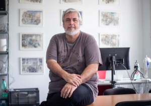 Bearded man with gray hair sits at a desk in a bright office, framed pictures on the wall behind him.