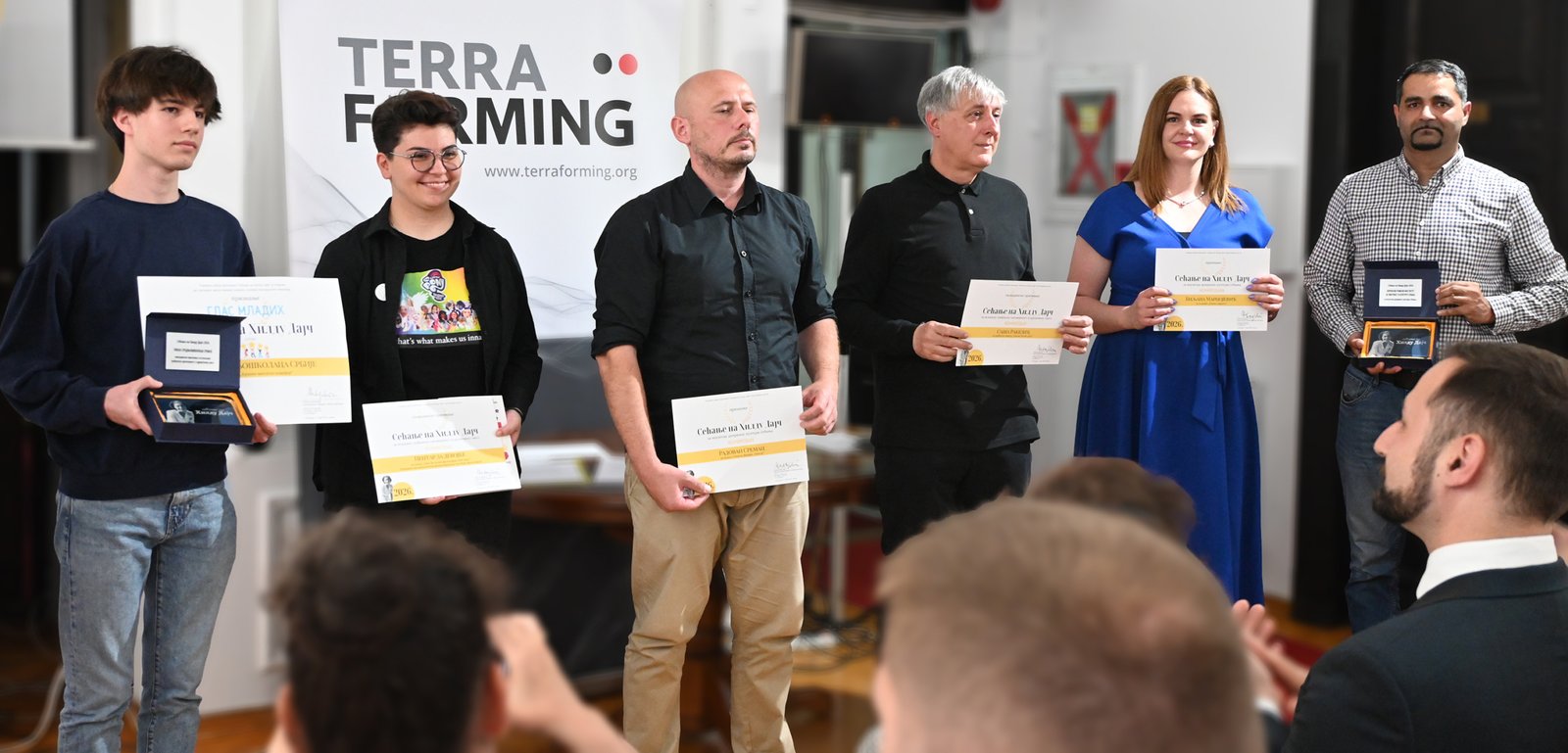 Six award recipients stand in a line holding certificates and plaques, with a Terra Forming banner in the background.