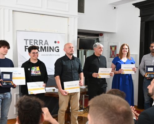 Six people stand in a row on a stage holding certificates, with a Terra Forming banner behind them and an audience in the foreground at a ceremony.