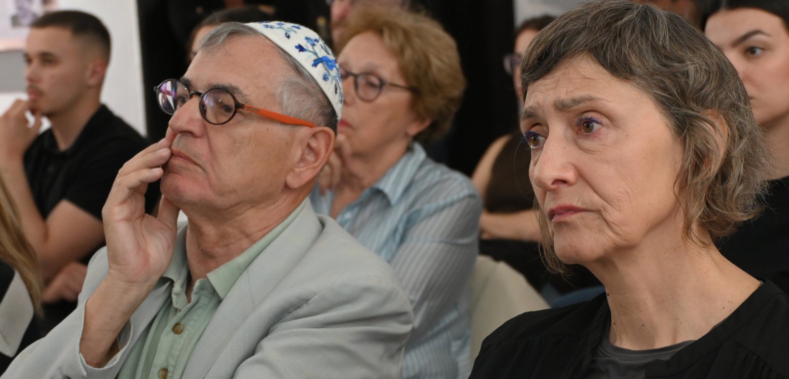 Older man with a patterned kippah and glasses rests his chin on his hand while listening at a conference; beside him, a serious-looking woman with short gray hair looks forward.