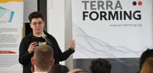 Speaker with round glasses presenting at a TerraForming event, speaking into a microphone while looking at a smartphone in hand.