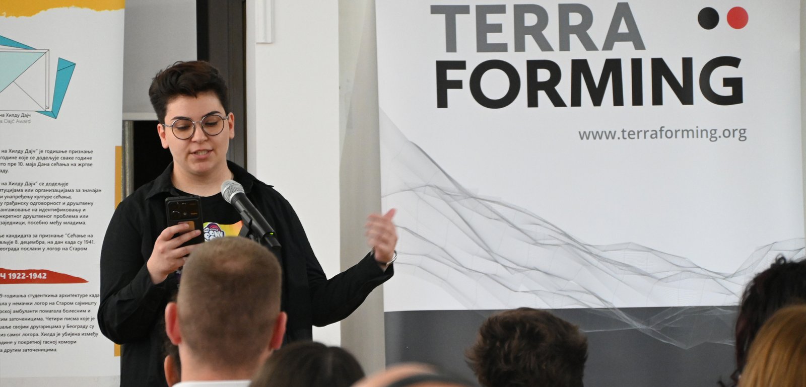 Speaker with round glasses presenting at a TerraForming event, speaking into a microphone while looking at a smartphone in hand.