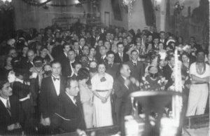 Black-and-white photo of a crowded indoor party with men in suits and women in dresses, facing the camera; foreground shows stage or equipment, suggesting a formal celebration.