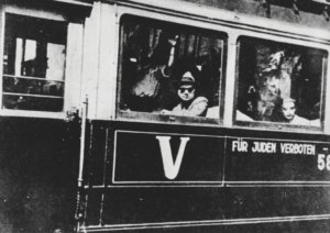 Black-and-white photo of a bus with passengers visible in the windows; on the side is a large 'V' and the German text 'Für Juden verboten' (for Jews prohibited).