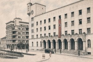 Historic street scene with a large government building displaying a swastika banner above arched entryways and a vintage car in front.