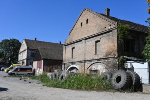Old brick industrial building with arched openings and overgrown vegetation, tires stacked along the wall in front.