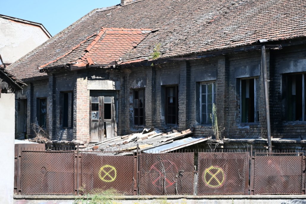 Abandoned brick building with a collapsed tiled roof, broken windows, and a rusty fence in the foreground.