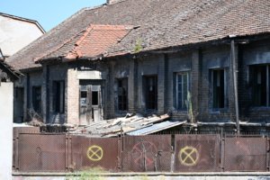 Abandoned brick building with a collapsed tiled roof, broken windows, and a rusty fence in the foreground.