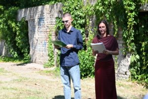 Two presenters stand outdoors beside a ivy-covered stone wall, reading from folders.