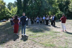 A group of people standing in a sunlit park, some holding sunflowers, listening to a speaker at the front.