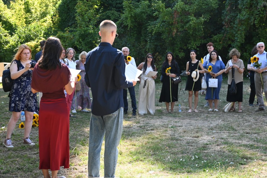 Group of people outdoors standing in a semicircle, some holding sunflowers, listening to a speaker facing away from the camera.