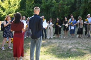Group of people outdoors standing in a semicircle, some holding sunflowers, listening to a speaker facing away from the camera.