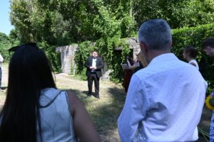 Man in a dark suit addressing an outdoor gathering beside a ivy-covered brick wall as people listen, some holding papers.