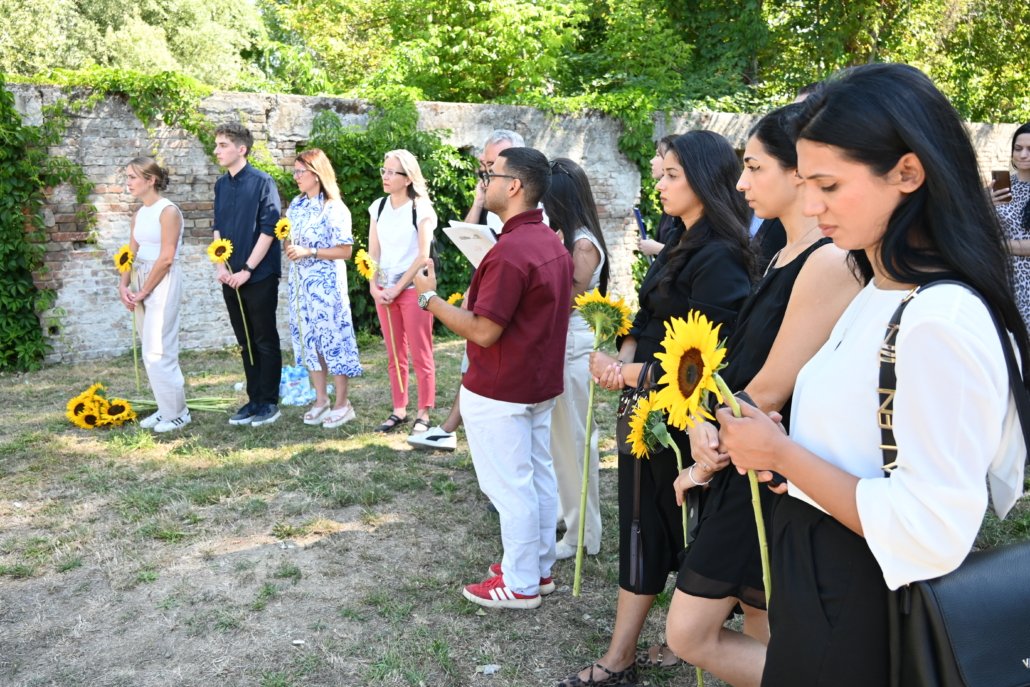 Line of diverse people standing outdoors in a row, each holding a sunflower, beside a brick wall and greenery.