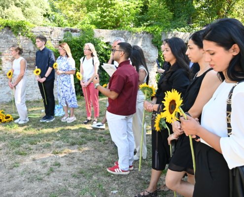 Line of diverse people standing outdoors in a row, each holding a sunflower, beside a brick wall and greenery.