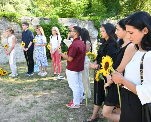 Line of diverse people standing outdoors in a row, each holding a sunflower, beside a brick wall and greenery.