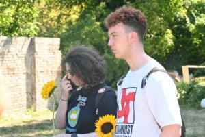 Two people outdoors with sunflowers; left person with curly hair holds a sunflower, right person wears a white shirt with a red graphic, greenery in background.