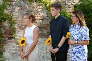 Three young adults stand side by side outdoors, each holding a sunflower in front of a brick wall with greenery behind them.