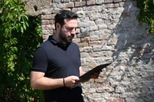 Bearded man in a navy polo shirt standing outdoors, looking at a clipboard against a brick wall.