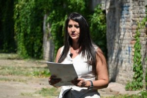 Woman with long black hair wearing a white sleeveless dress reads from a paper outdoors beside a brick wall and greenery.
