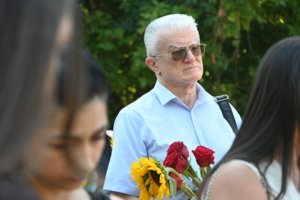 Older man with white hair and sunglasses in a light blue shirt holding a bouquet of red roses and sunflowers.