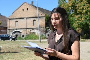 Young woman with long dark hair reads a document outdoors, in front of a brick building on a sunny day.