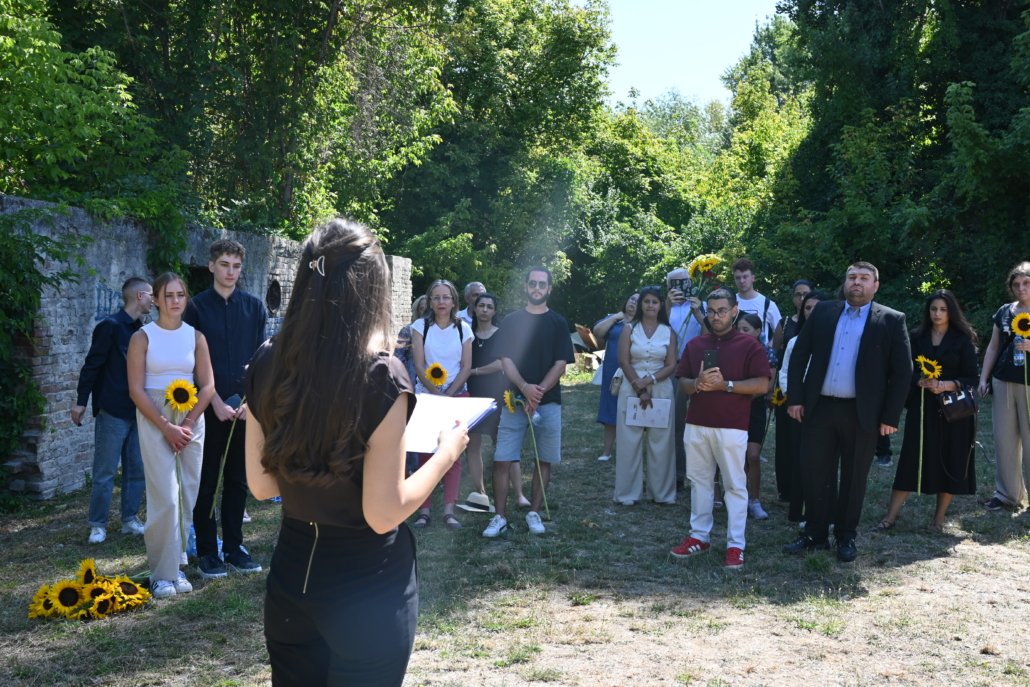 Speaker facing a diverse crowd outdoors, several attendees hold sunflowers near a stone wall.