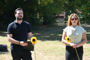 Two adults stand in a sunny park, each holding a sunflower; man on the left has a tablet, woman on the right wears sunglasses.