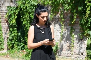 Woman in a black sleeveless dress outdoors, looking at her phone with a vine-covered brick wall behind her, sunlit scene.