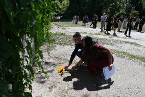 Two people kneel on a cracked pavement placing yellow sunflowers; a line of guests stands in the background holding sunflowers outdoors.