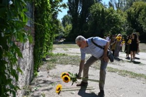 Man bending to pick sunflowers from the ground as a group of people holding sunflowers stand behind him outdoors near an ivy-covered brick wall.