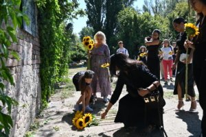 Group of people outdoors holding sunflowers, some bending to arrange flowers along a path by a brick wall and green foliage.