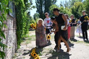 Group of people outdoors placing bright sunflower stems along a brick wall, some kneeling to lay flowers in a memorial gesture.