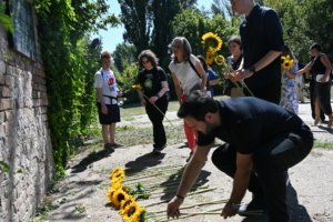Group of people outdoors placing bright yellow sunflowers on the ground beside a brick wall during a sunny gathering, likely a memorial.