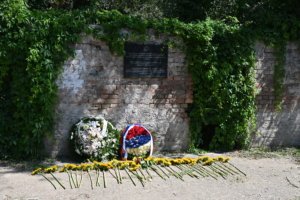 Brick wall with ivy and a dark memorial plaque, floral wreaths arranged on the ground in front.