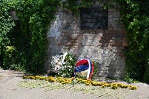 Brick wall partly covered with ivy, with a dark memorial plaque mounted on it; in front are floral wreaths and long-stemmed yellow flowers laid on the ground, left to right.