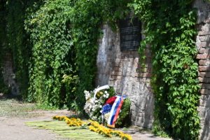 Brick wall covered in dense ivy with a dark memorial plaque and floral wreaths at the base. (Memorial site)