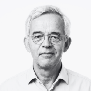 Portrait of an older man with white hair and round glasses wearing a light shirt, looking at the camera.