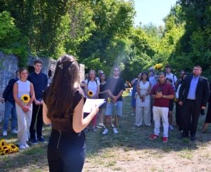 Presenter with notebook addressing a crowd outdoors; several attendees hold sunflowers.