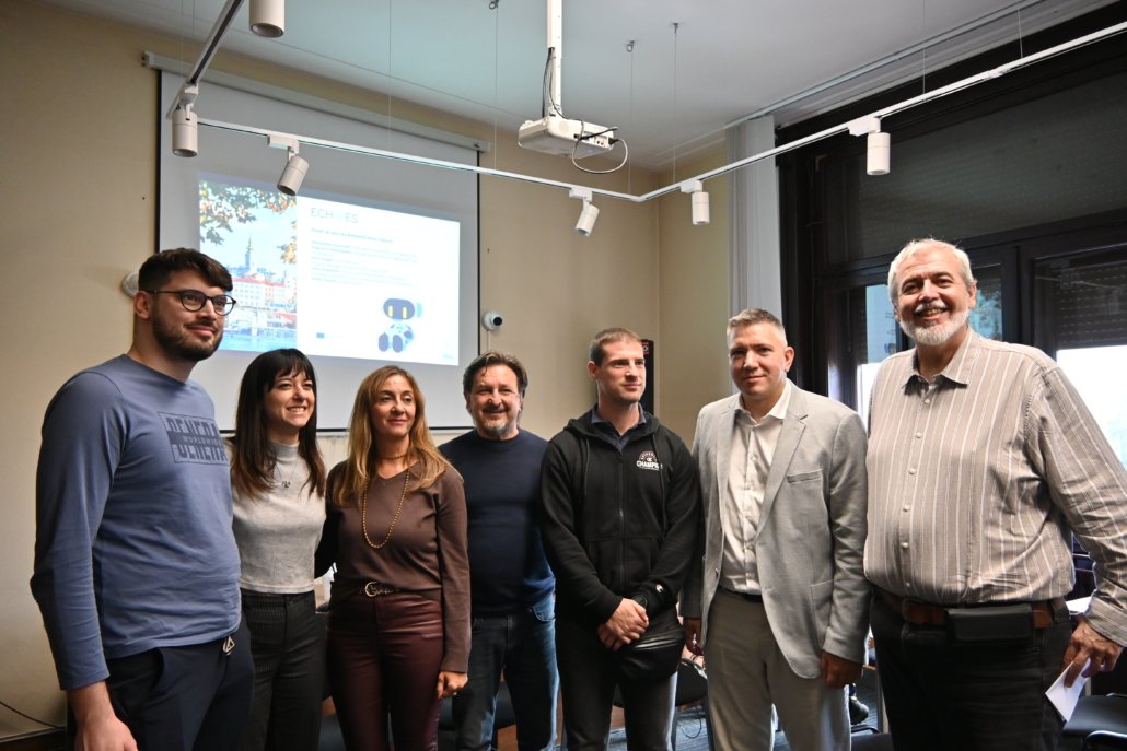 Group of eight adults posing together in a meeting room, projector screen and window behind them.