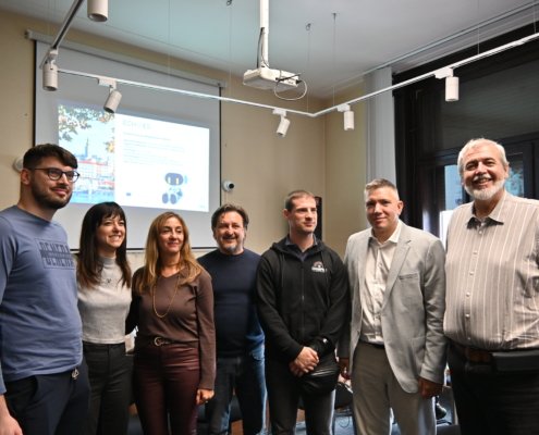 Group of eight adults posing together in a meeting room, projector screen and window behind them.